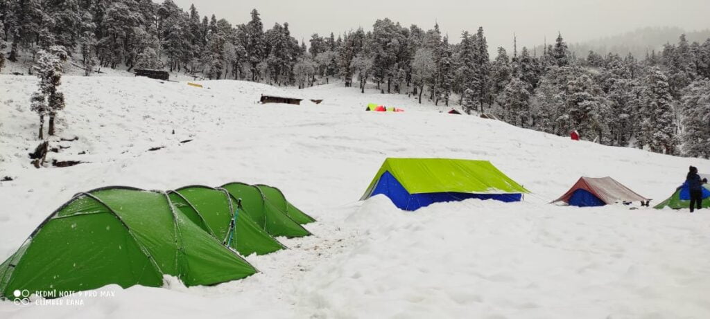 Snow campsite with green trekking tents during the Kedarkantha trek in Uttarakhand