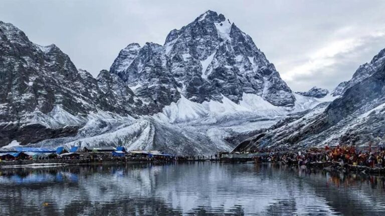Pilgrims and trekkers gathered around Manimahesh Lake with snow-covered mountains reflected in the water.
