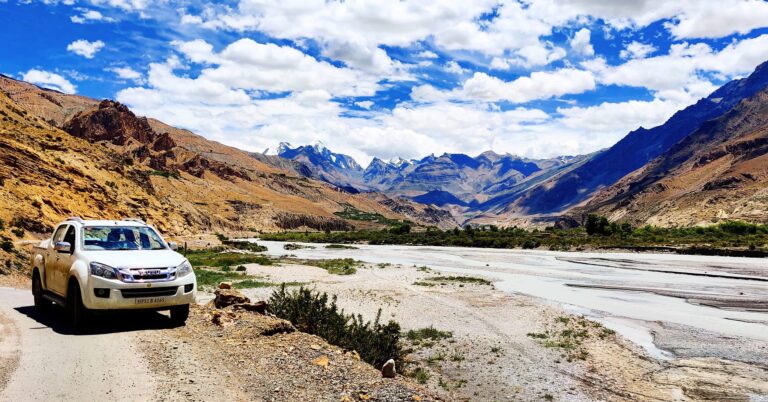 SUV driving along a scenic river valley road in Spiti Valley, Himachal Pradesh, surrounded by high-altitude cold desert mountains