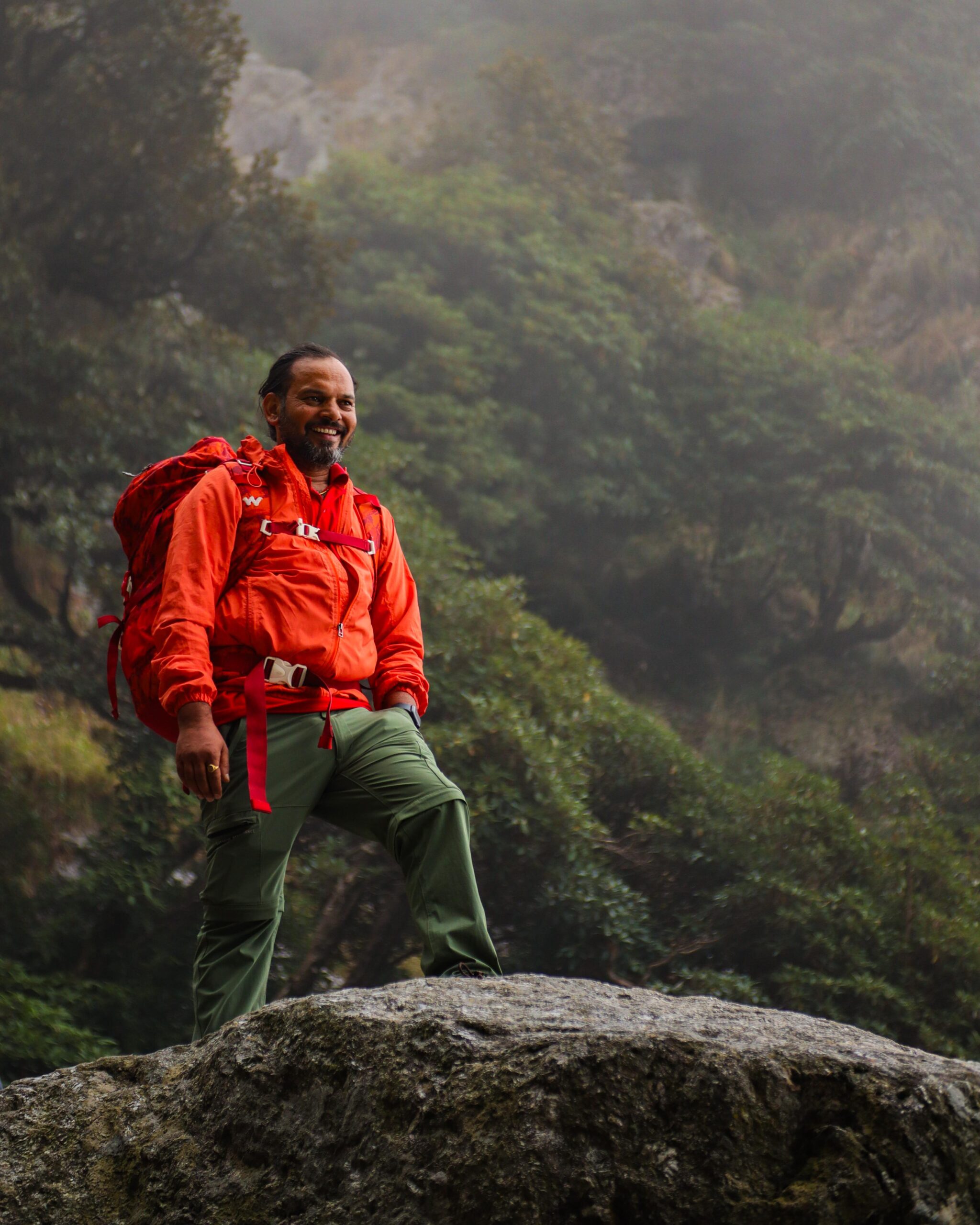 Smiling male trekker in red hiking gear standing in front of a forested mountain backdrop.