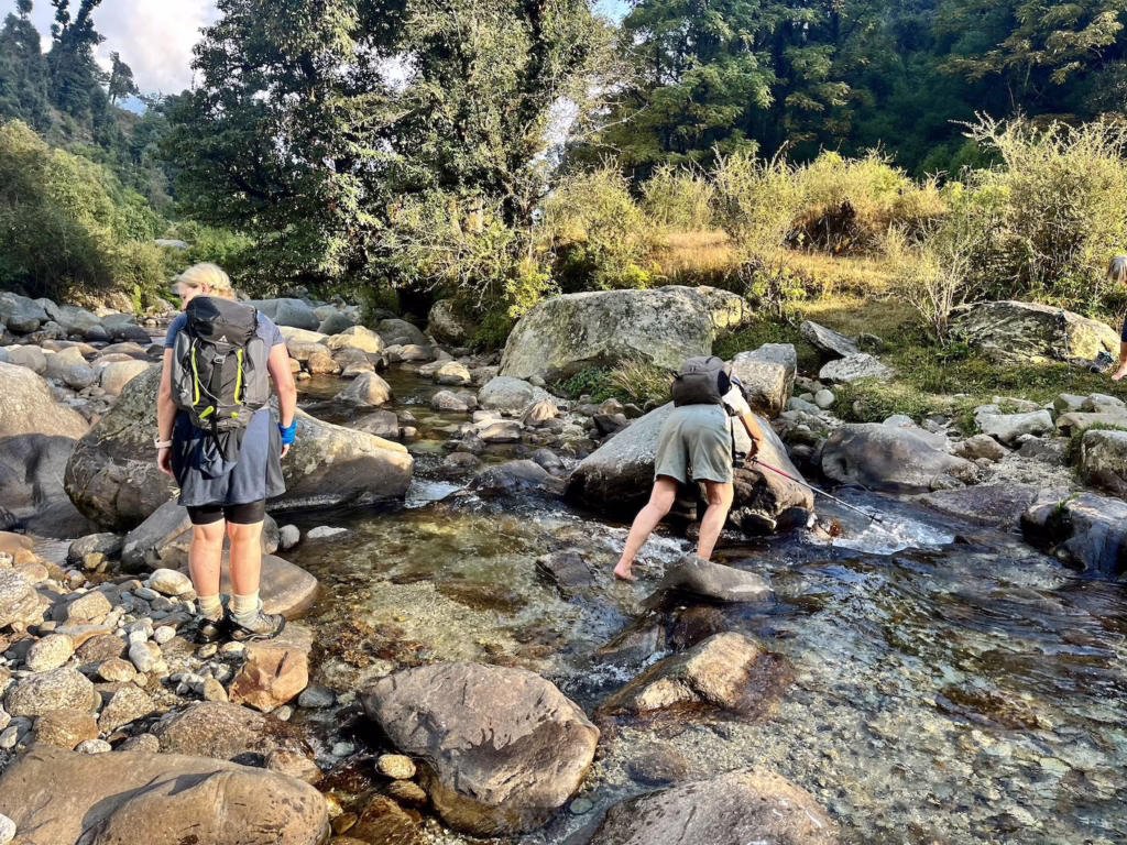 🌿 Himalayas on the Edge: How to Trek Without Leaving a Scar 6 Two trekkers cross a shallow rocky stream in the Himalayas surrounded by forest and golden afternoon light.