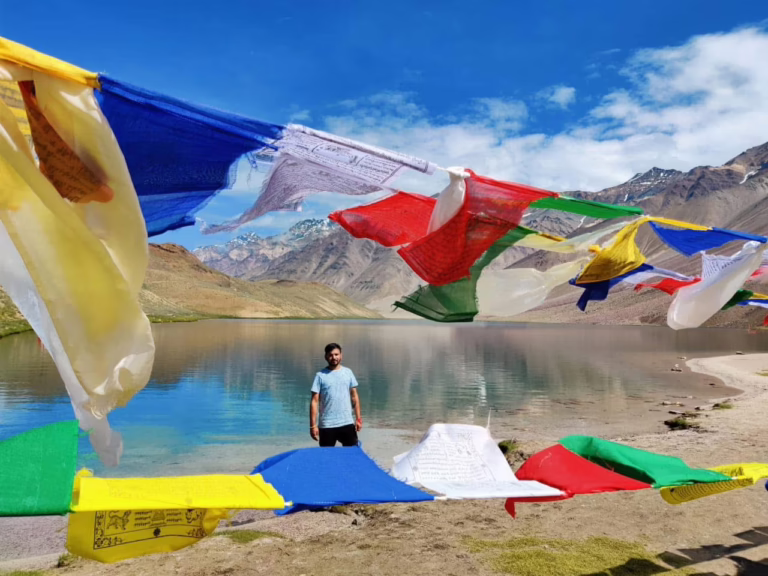 Prayer flags at Chandratal Lake with a man standing by the shore in Spiti Valley, Himachal Pradesh.