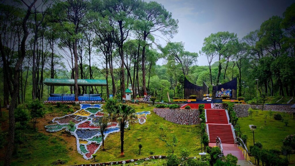 A landscaped garden in Dharamshala with a large floral map of India, red stairways, and a memorial stage surrounded by tall pine trees.