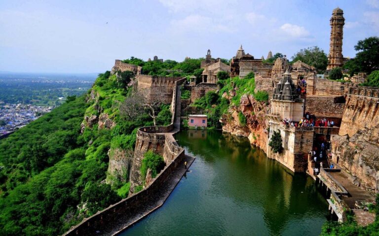 Aerial view of Chittorgarh Fort in Rajasthan with ancient stone walls, temples, and water reservoir surrounded by lush green hills.