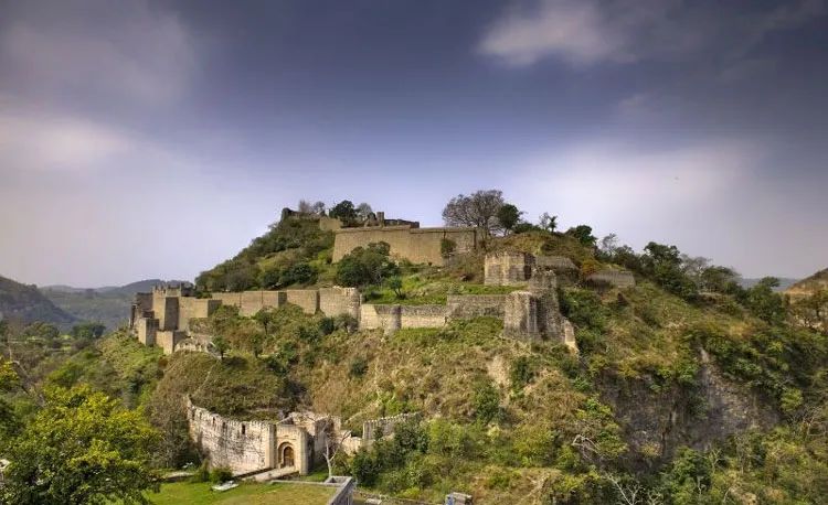 Ancient Kangra Fort on a green hill surrounded by trees under a cloudy sky in Himachal Pradesh, India.