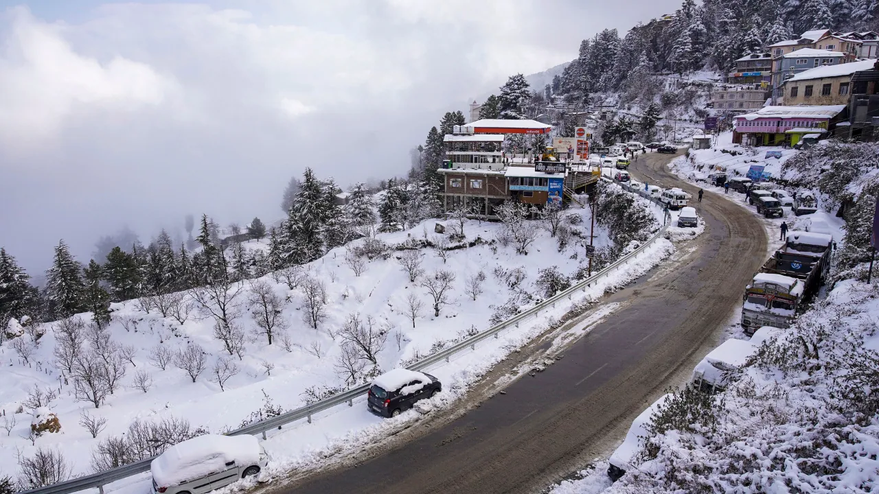 Snow-covered road in Kufri with hillside buildings, parked cars, and pine trees during winter in Himachal Pradesh.