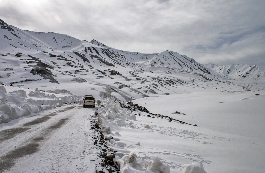 Car driving on a snow-covered mountain road surrounded by rugged snowy peaks under a cloudy winter sky in Ladakh.
