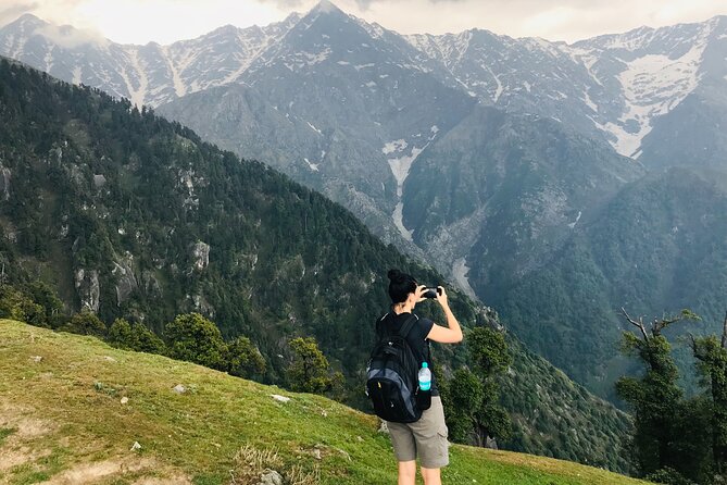 Solo trekker with a backpack photographing snow-capped Himalayan mountains from a grassy ridge, capturing the spirit of first-time mountain exploration and adventure