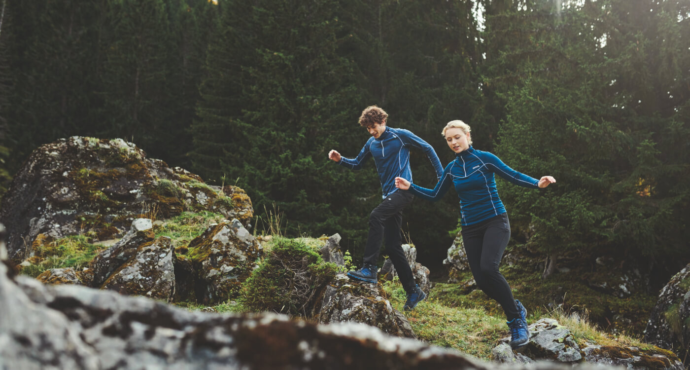 Couple wearing thermal base layer clothing while hiking through a forested mountain trail, demonstrating proper layering for cold-weather mountain camping and trekking