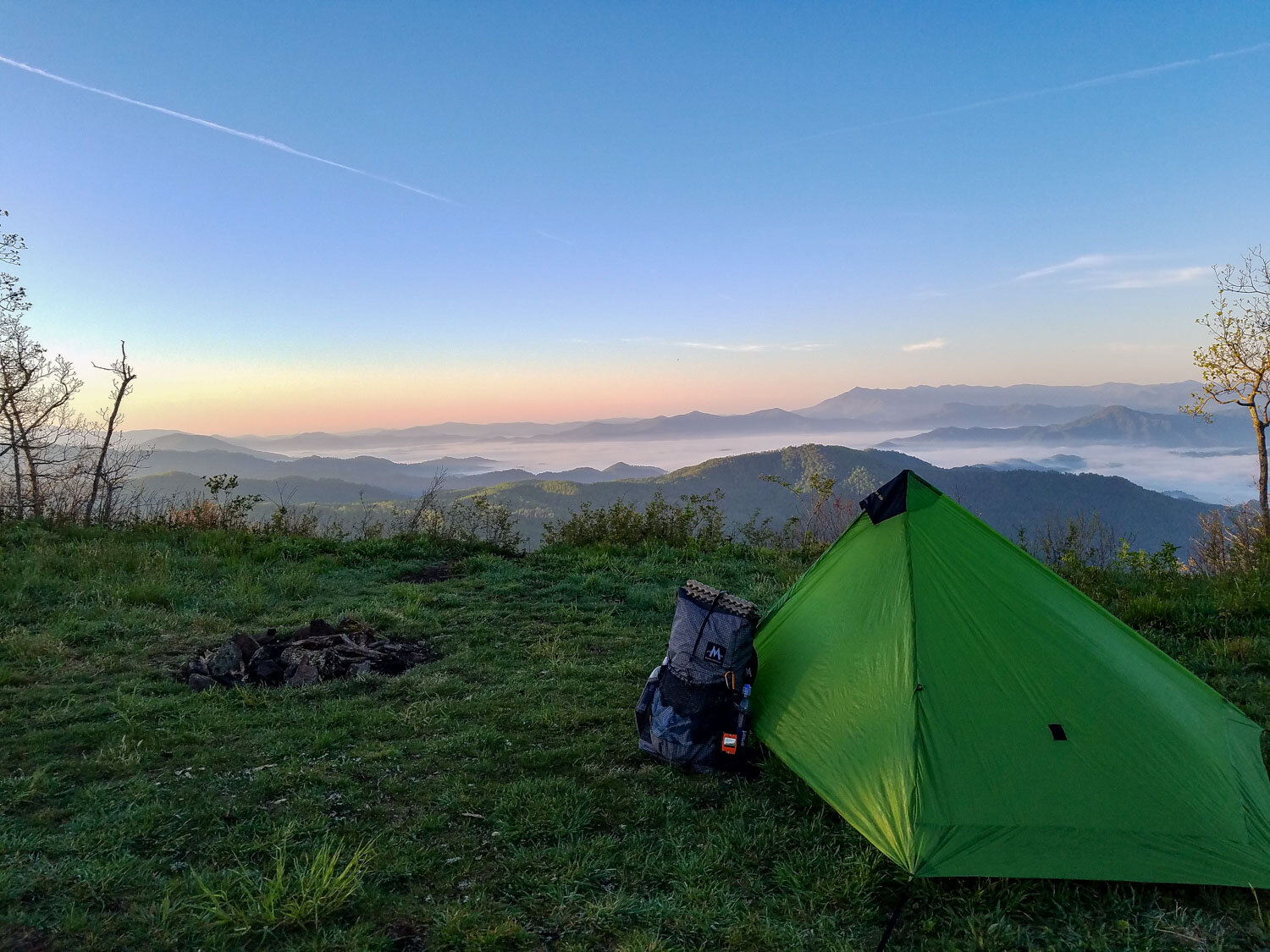 Green camping tent pitched on a grassy mountain ridge at sunrise with layered Himalayan hills and mist-filled valleys in the background
