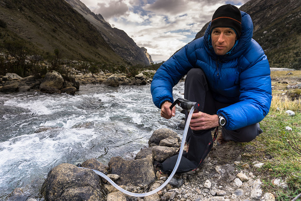 Man using a portable water filtration system to collect drinking water from a mountain stream during high-altitude camping and trekking