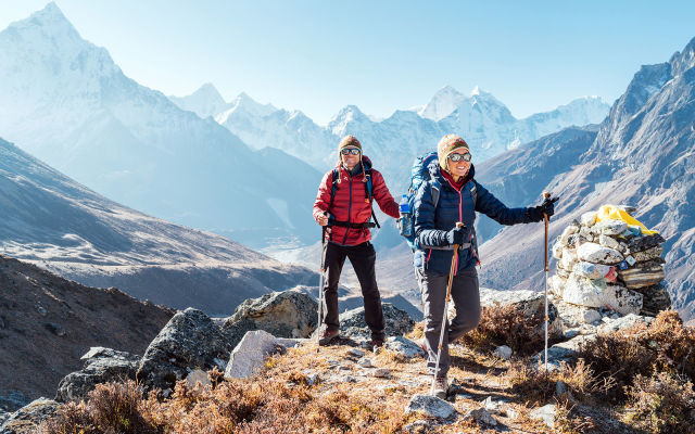 Two trekkers walking together on a Himalayan mountain trail near a stone cairn, with snow-covered peaks in the background, representing guided group trekking experiences for solo travelers in India.